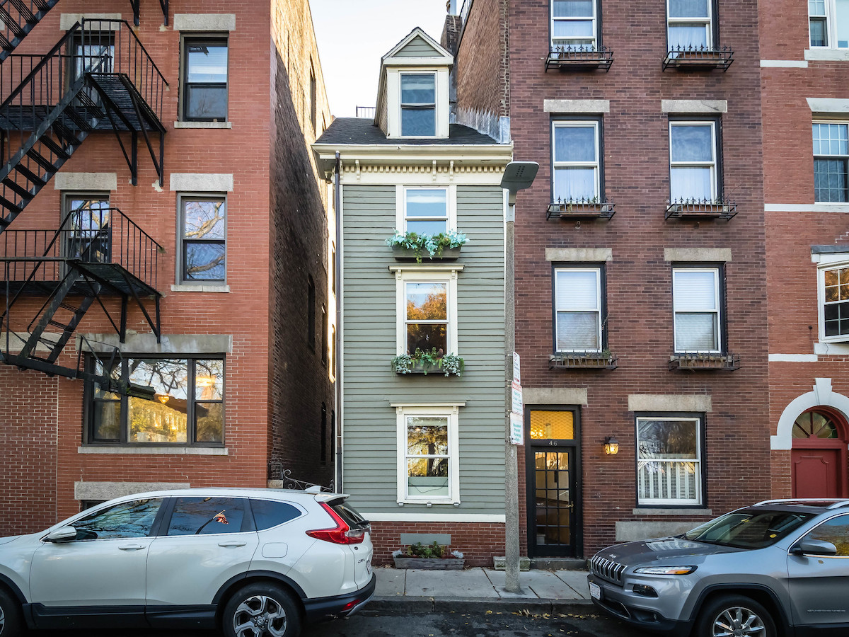 A skinny, three level house painted green with flowers spilling out of window boxes. The house is sandwiched in between two normal sized brick buildings. A house so skinny it's a true Boston hidden gem
