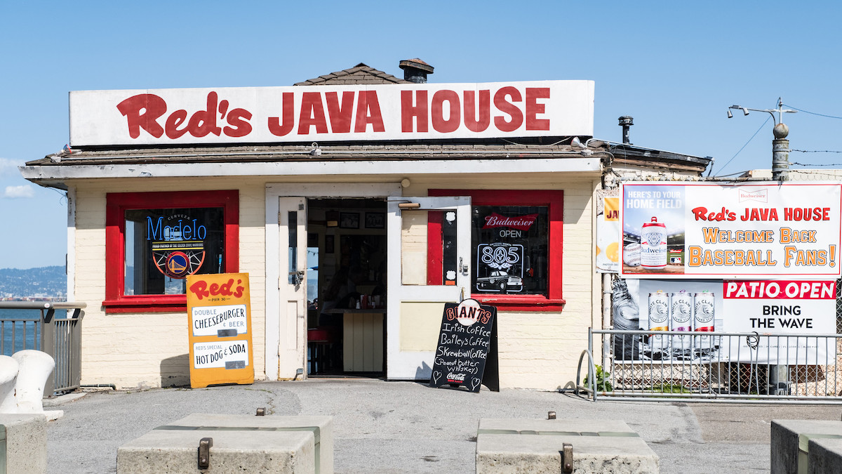 exterior shot of a small white building on a pier with a blue sky. Big red letters spell "Red's JAVA HOUSE" on a sign