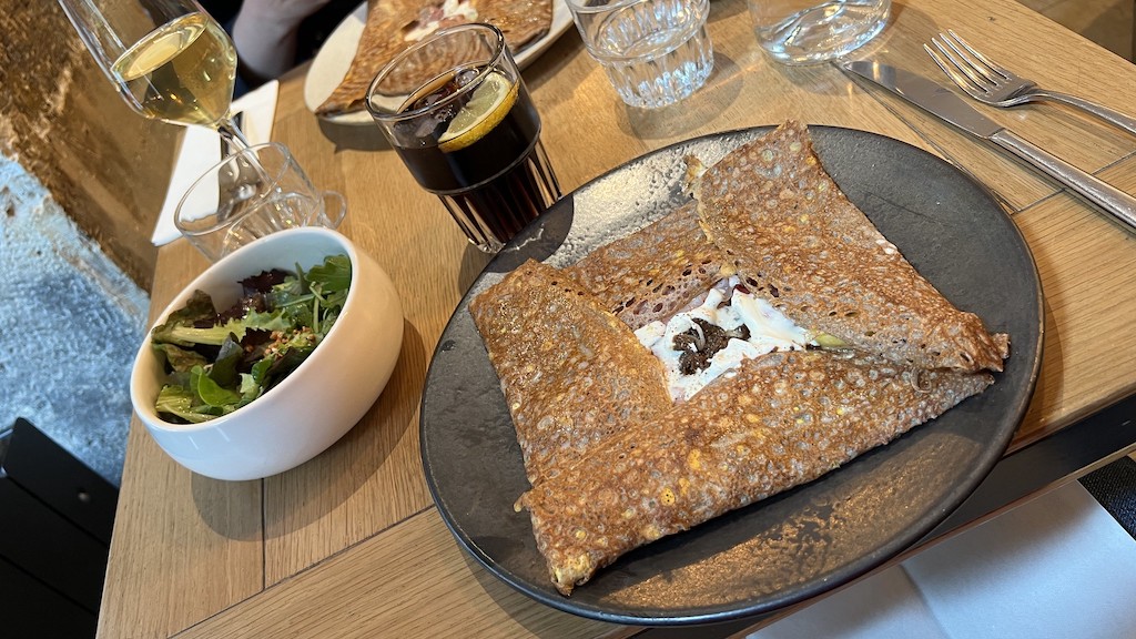 Square, folded buckwheat crepe with cheesey filling on parisian restaurant table with salad and a soft drink