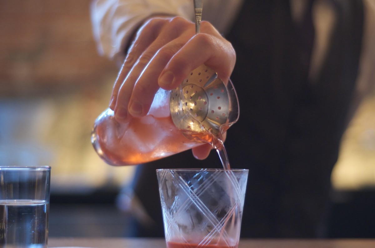 A bartender pours a pale red cocktail from a shaker into a cocktail glass.