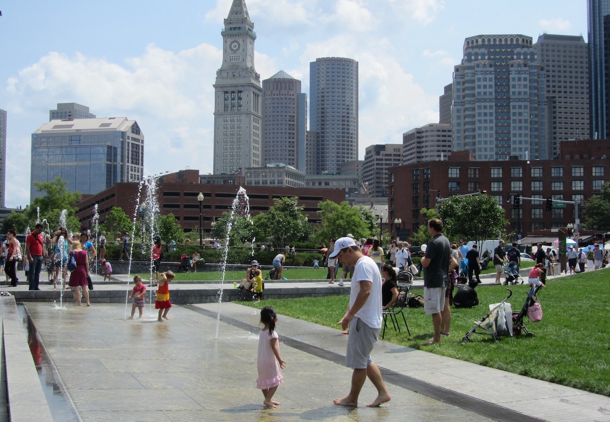 children play in water fountains and families sit in the grass of the Rose Kennedy Greenway in Boston