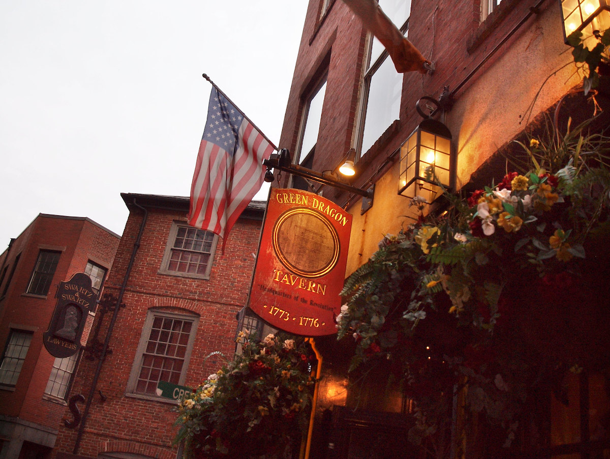 An outdoor shot of a hanging wooden sign on a brick building with the American flag flying in the background. The sign is painted red and reads, "Green Dragon Tavern"