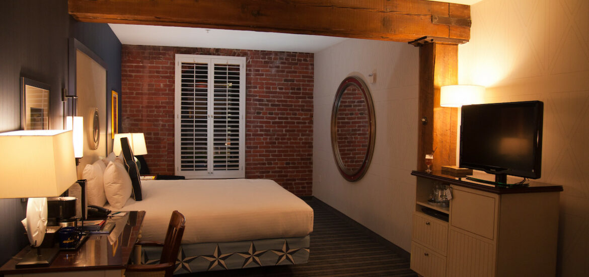 Interior of a hotel room with walls painted white, an exposed brick wall with a window, and wooden beams on the ceiling