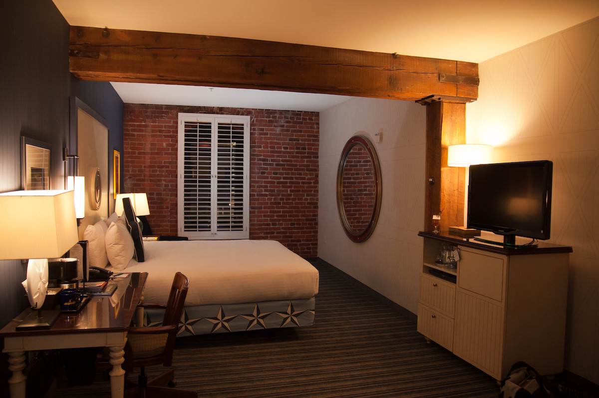 Interior of a hotel room with walls painted white, an exposed brick wall with a window, and wooden beams on the ceiling 