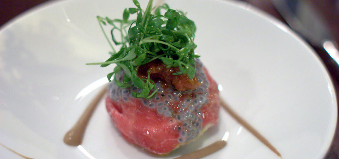 Extreme close up of a white place with a small piece of raw beef carpaccio, topped with greens