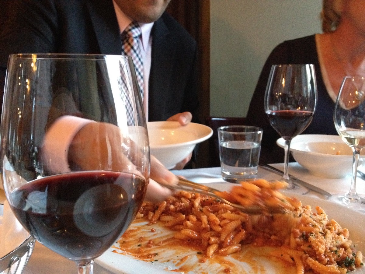 Two people sit at a table with red wine in wine glasses, as they share a plate of pasta