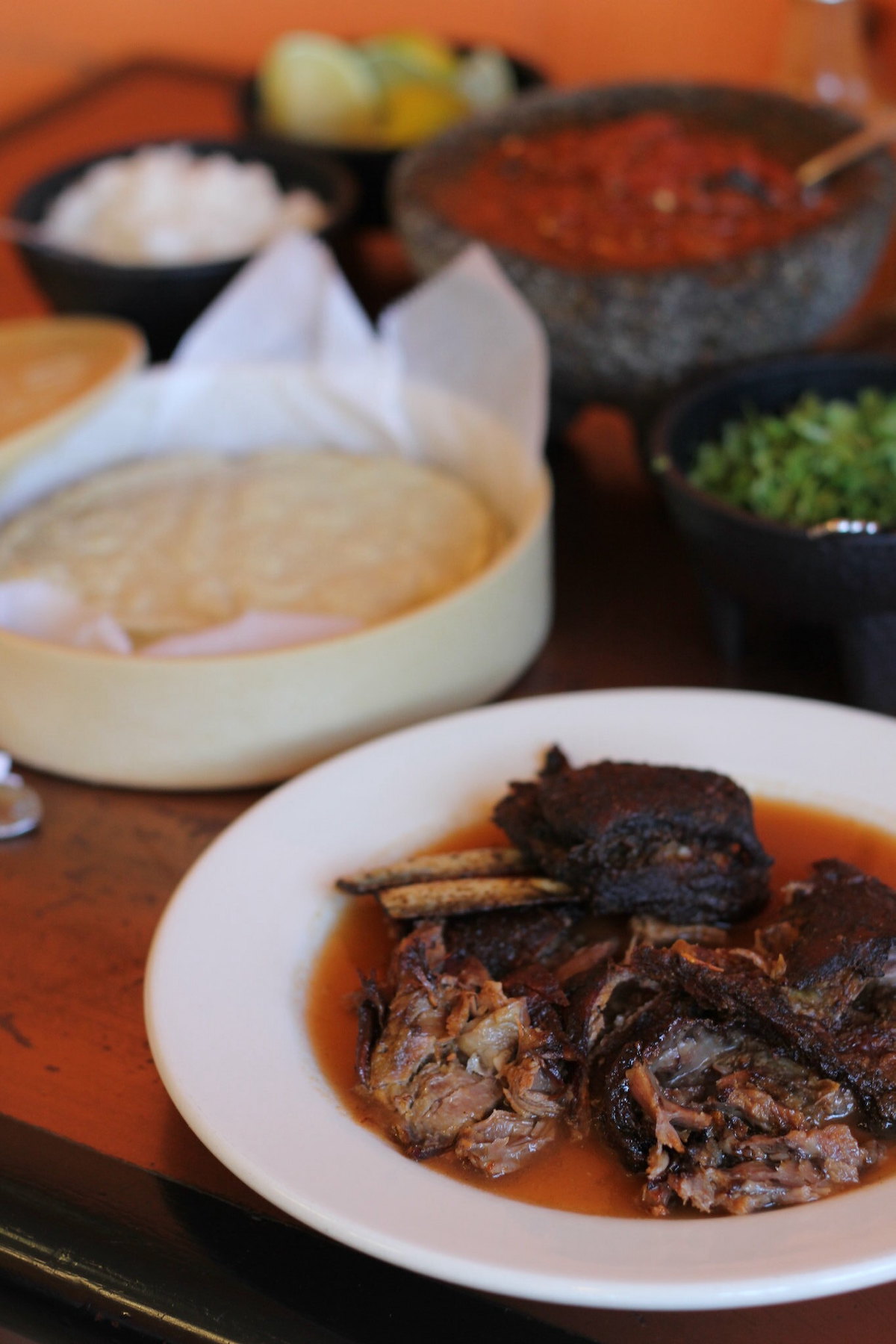 Close up of a table at Birrieria Zaragoza in Chicago. Stewed goat meat is on a white plate in the foreground, wtih salsa, tortillas, and toppings in the background