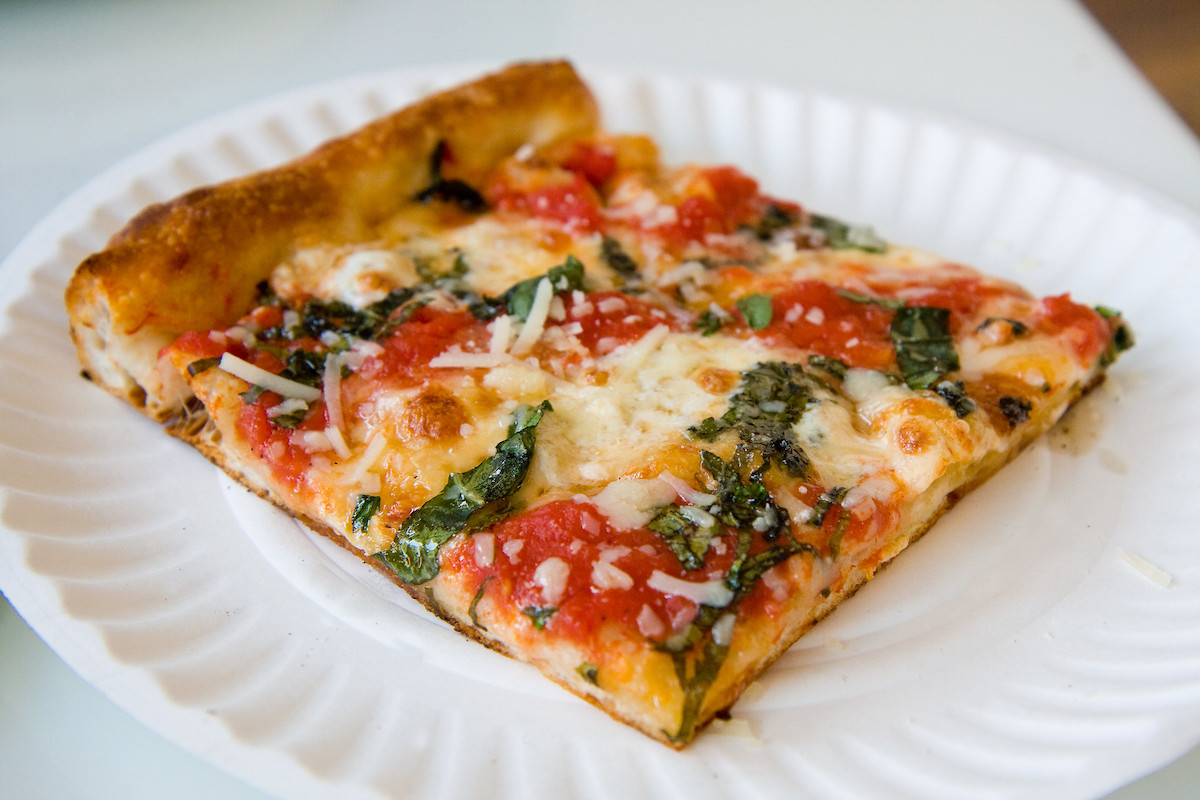 Close up of a white plate with a grandma slice of pizza. It is a thin crust in the shape of a square, topped with tomatoes, cheese, and basil