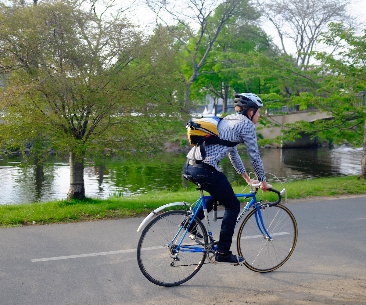 A person wearing jeans, a blue button up shirt, a cross-body bag, and a helmet rides a bicycle along the Charles River in Boston