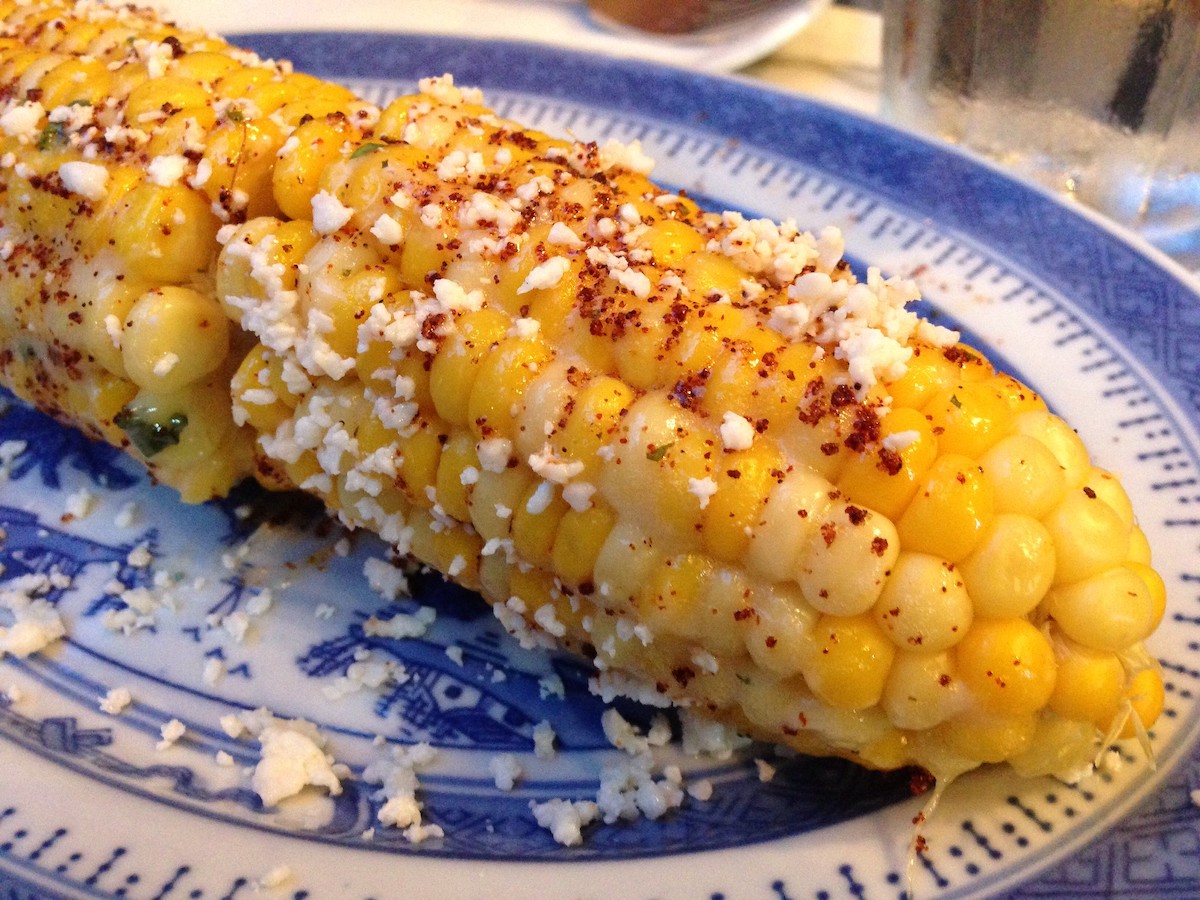 extreme close up of Mexican street corn on a blue and white plate. The yellow corn is dusted with red seasoning and crumbly white cheese.