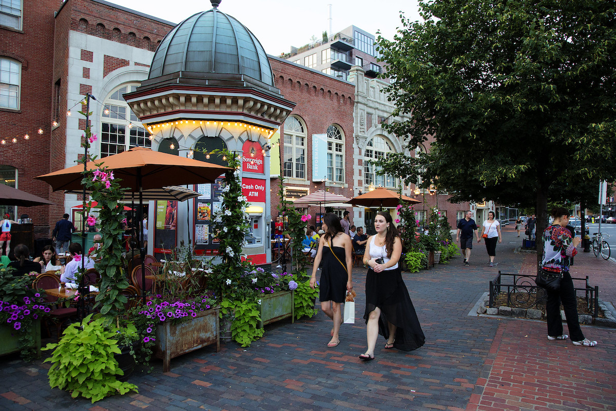 Two women walk down a brick paved sidewalk past a restaurant's outdoor seating area with surrounded by verdant green plants