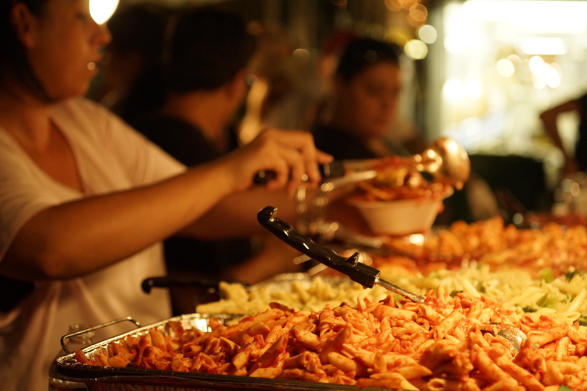 Close up of a tray of ziti with people serving up plates of pasta in the background. Fisherman's Feast is the oldest continuous Italian festival in Boston's North End.