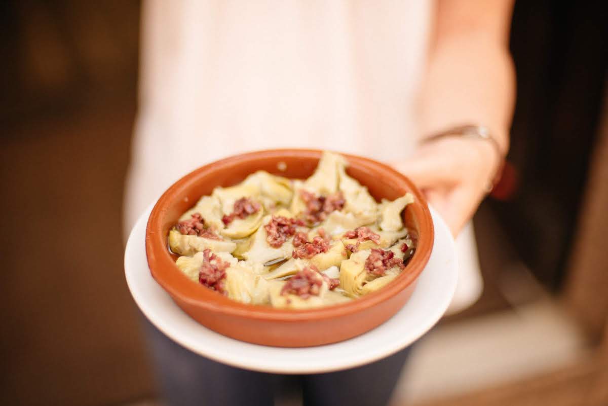 A person's hand holding a clay dish of artichoke hearts sprinkled with bits of Iberian ham.