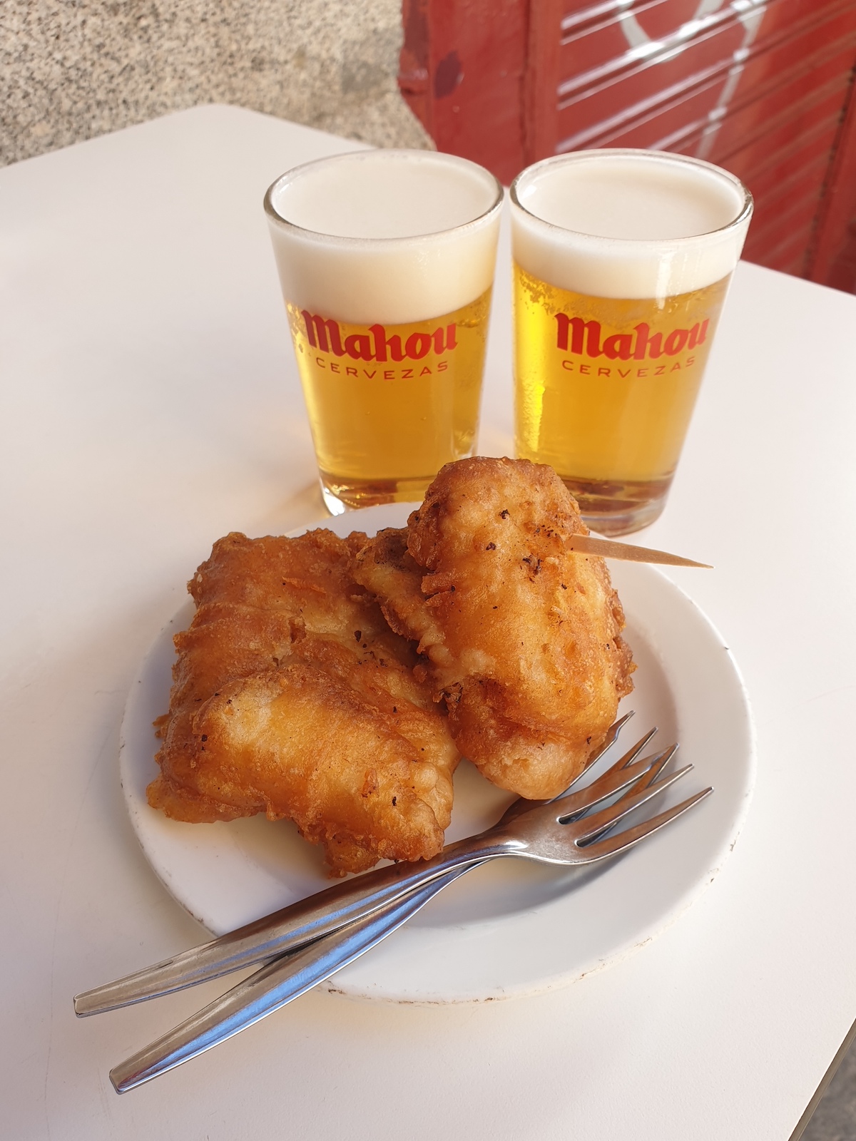 Pieces of fried codfish on a white plate in front of two small glasses of beer.