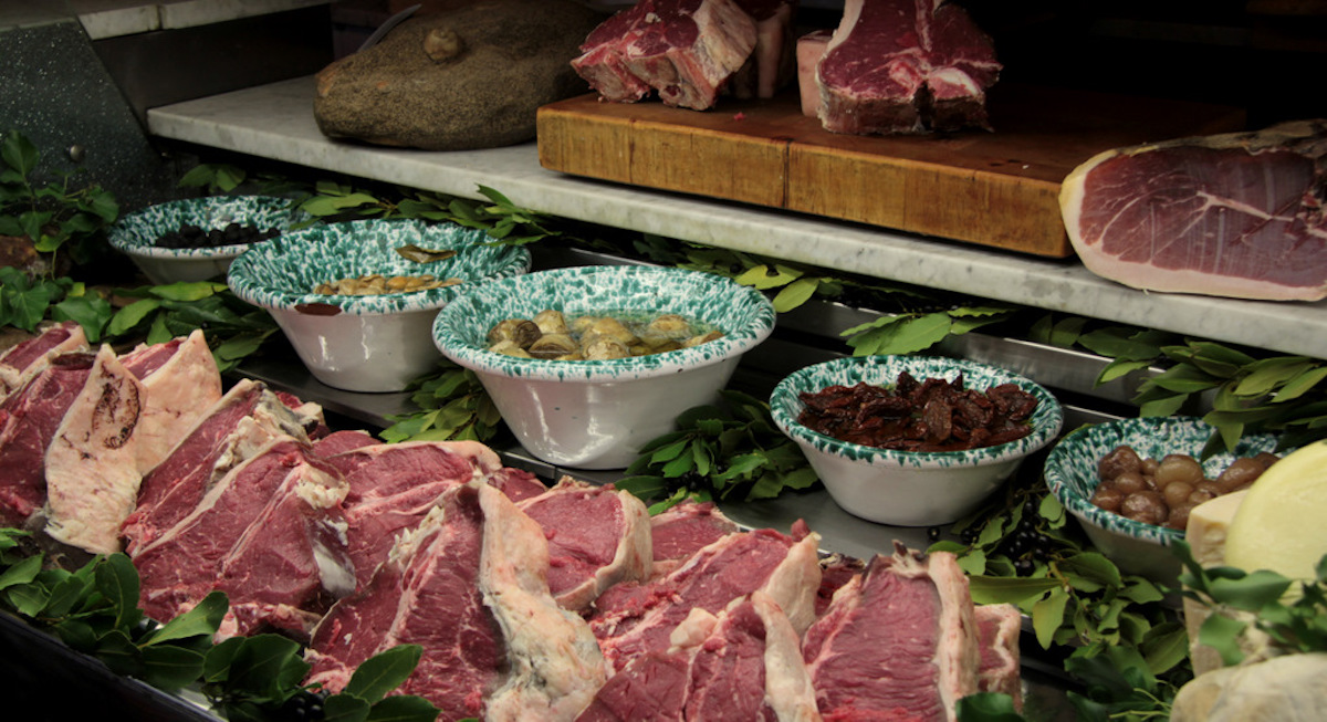 Raw steaks in a restaurant case with bowls of garnishes on the shelf above