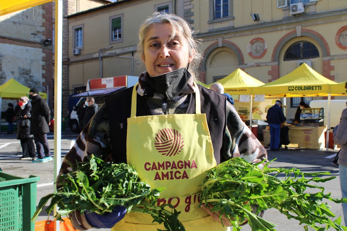 A woman in a yellow apron reading Campagna Amica smiling and holding two bunches of leafy greens