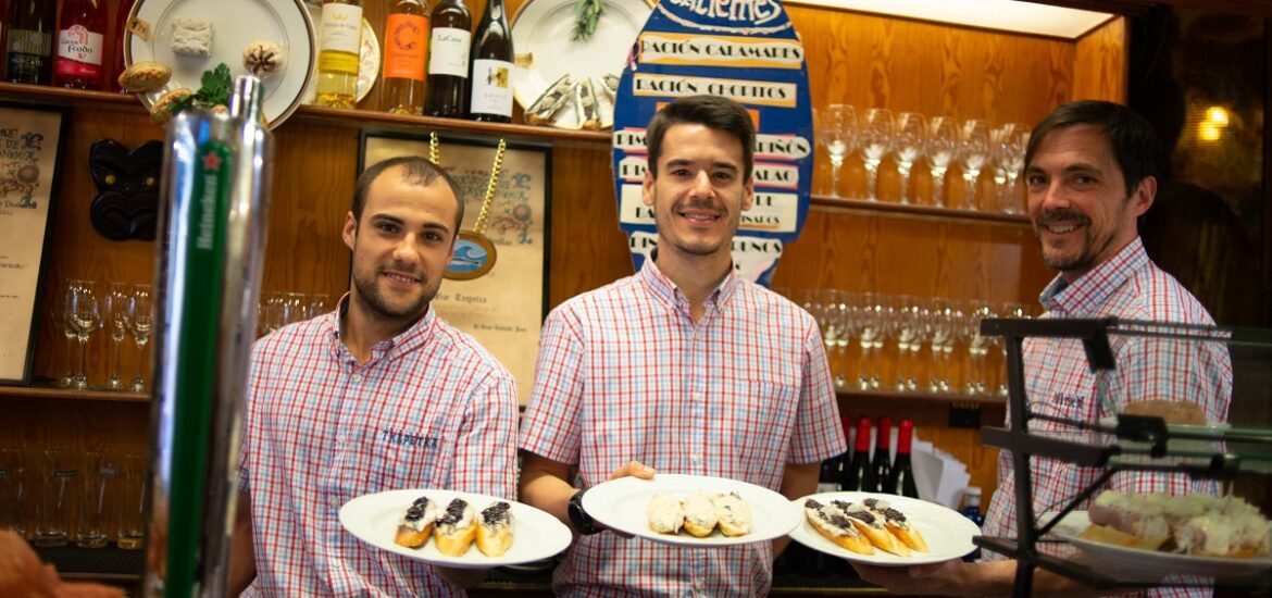 Three smiling servers at a Basque bar holding plates of pintxos at Bar Txapetxa.