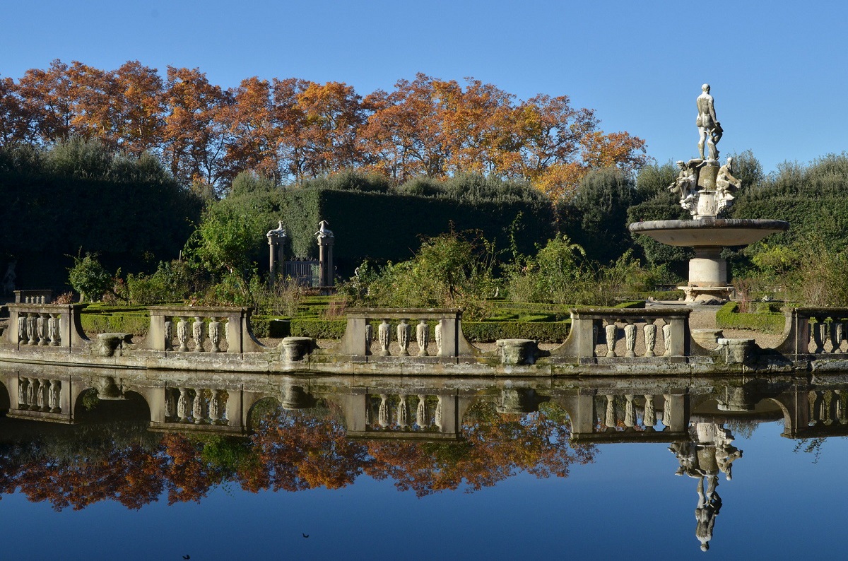 A large pool surrounded by stone statues and columns with trees in autumnal colors