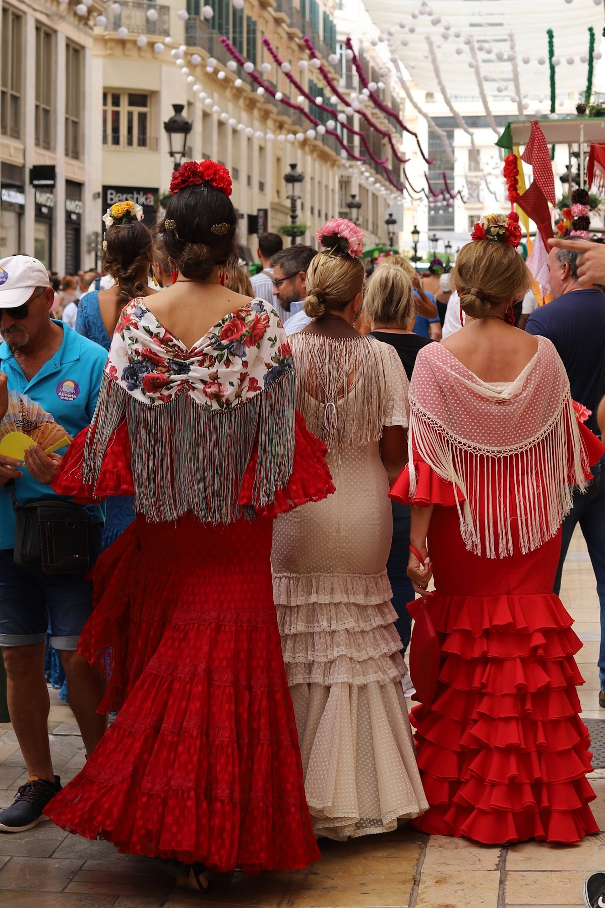 Shot following three women dressed in flamenco dresses down a street decorated with paper balls for feria