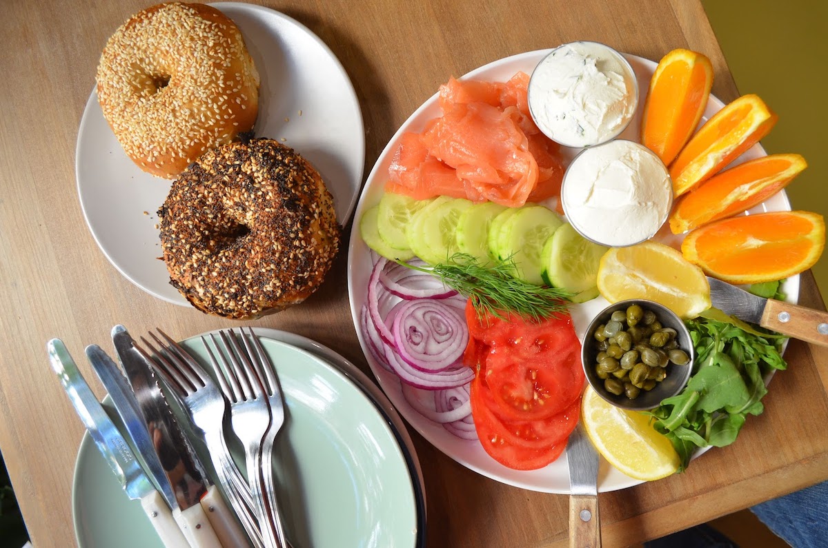 Two bagels on a small plate next to a larger plate with toppings such as salmon, cream cheese, and vegetables