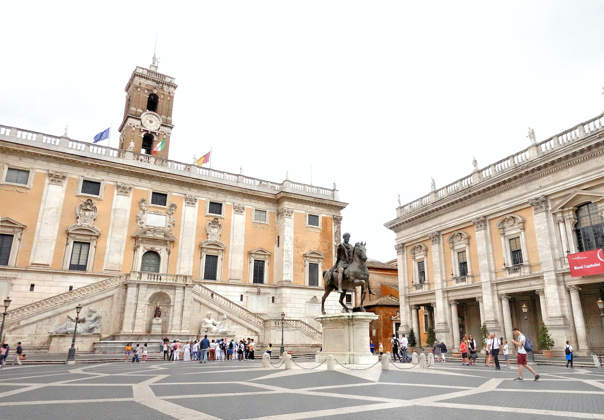 Two large stone buildings in a Roman piazza with a statue of a man on horseback in the center of the square.