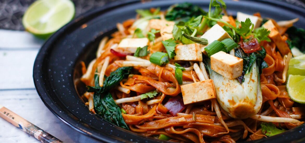 Plate of pad thai with noodles, tofu, lime slices, and cilantro with chopsticks