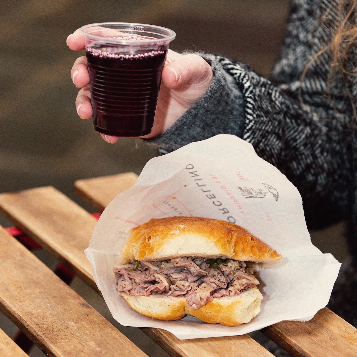 Lampredotto (tripe sandwich) in paper wrapping on a wooden table with a person's hand holding a plastic cup of red wine above