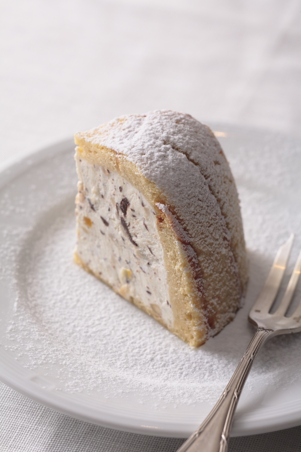 A slice of round, dome-shaped cake with filling and icing sugar with a fork on a plate