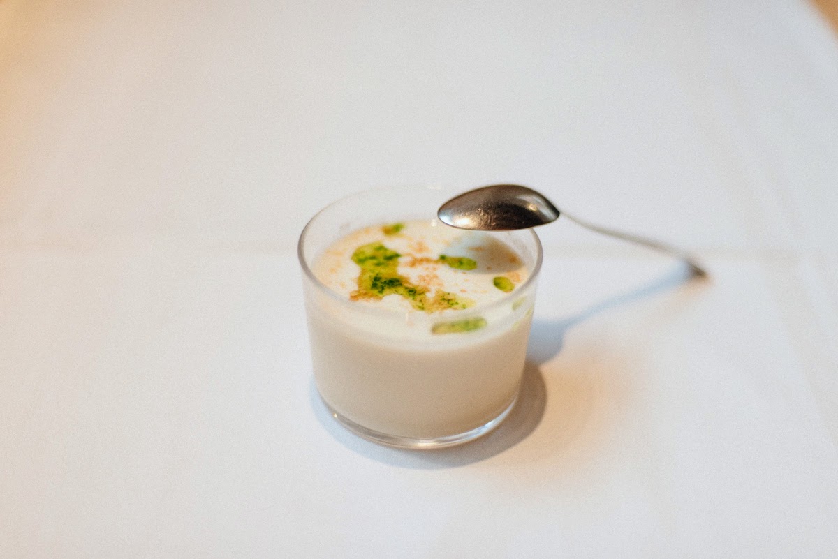 A small clear dish of chilled garlic soup garnished with grapes, with a metal spoon leaning against it.