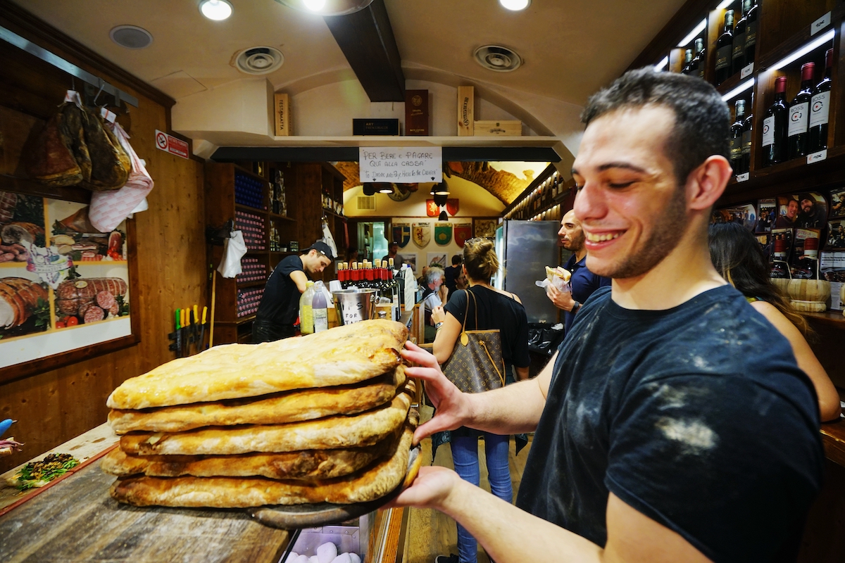 A man smiling while holding up a stack of focaccia-style bread loaves atop a restaurant counter