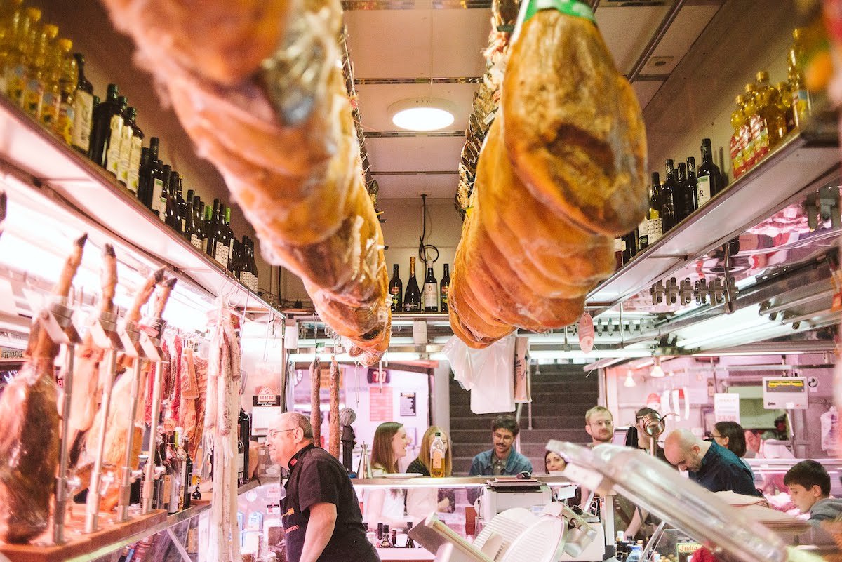 A crowded market stall in Madrid with cured ham legs hanging from the ceiling above.