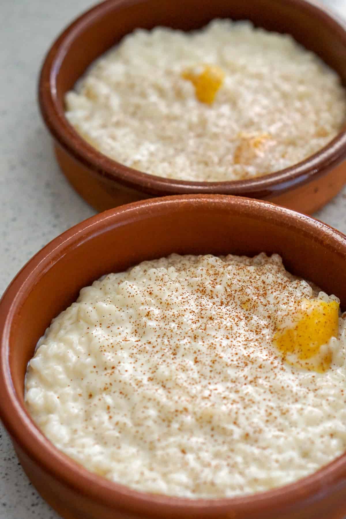 Two small clay dishes of rice pudding topped with cinnamon and a lemon peel