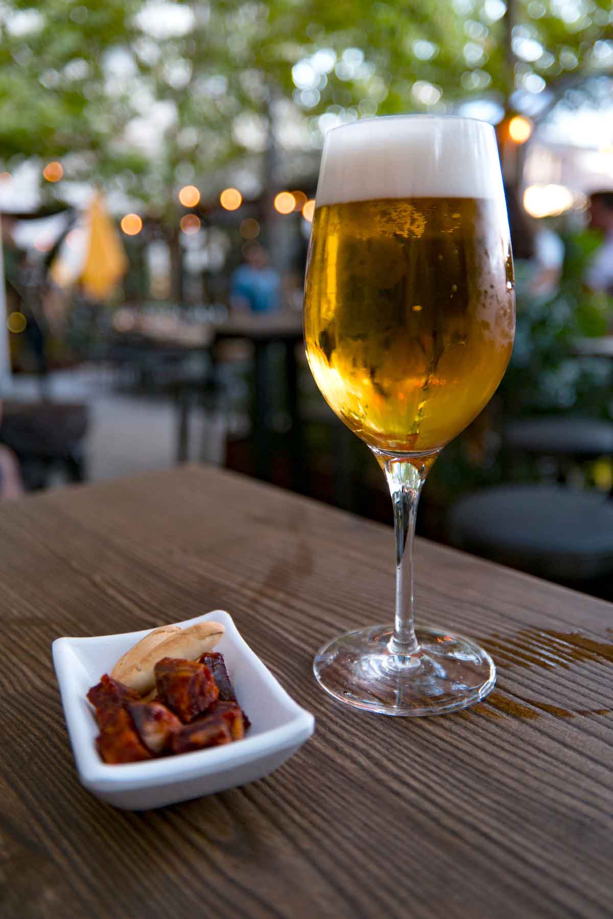 A tall glass of beer beside a small white dish of chorizo pieces and crunchy breadstick crackers on a wooden tabletop in an outdoor space