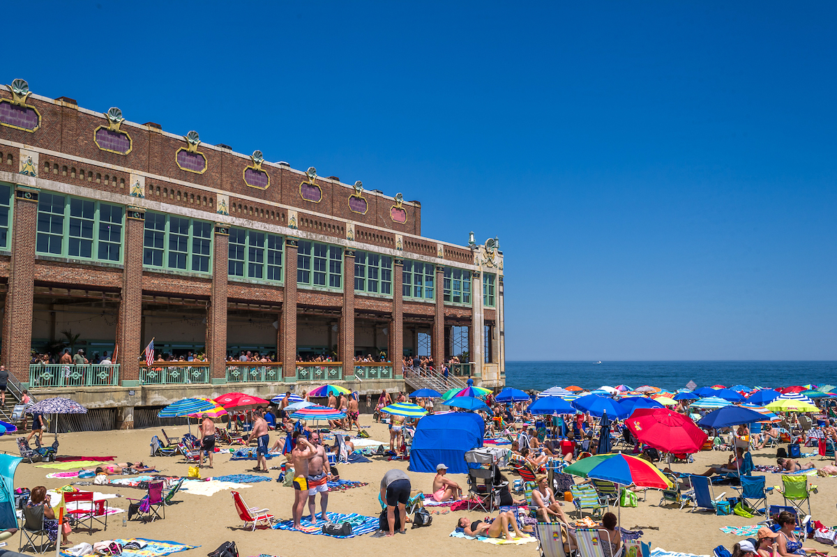 Busy beach full of people and umbrellas on a clear day with a large brick building at rear left