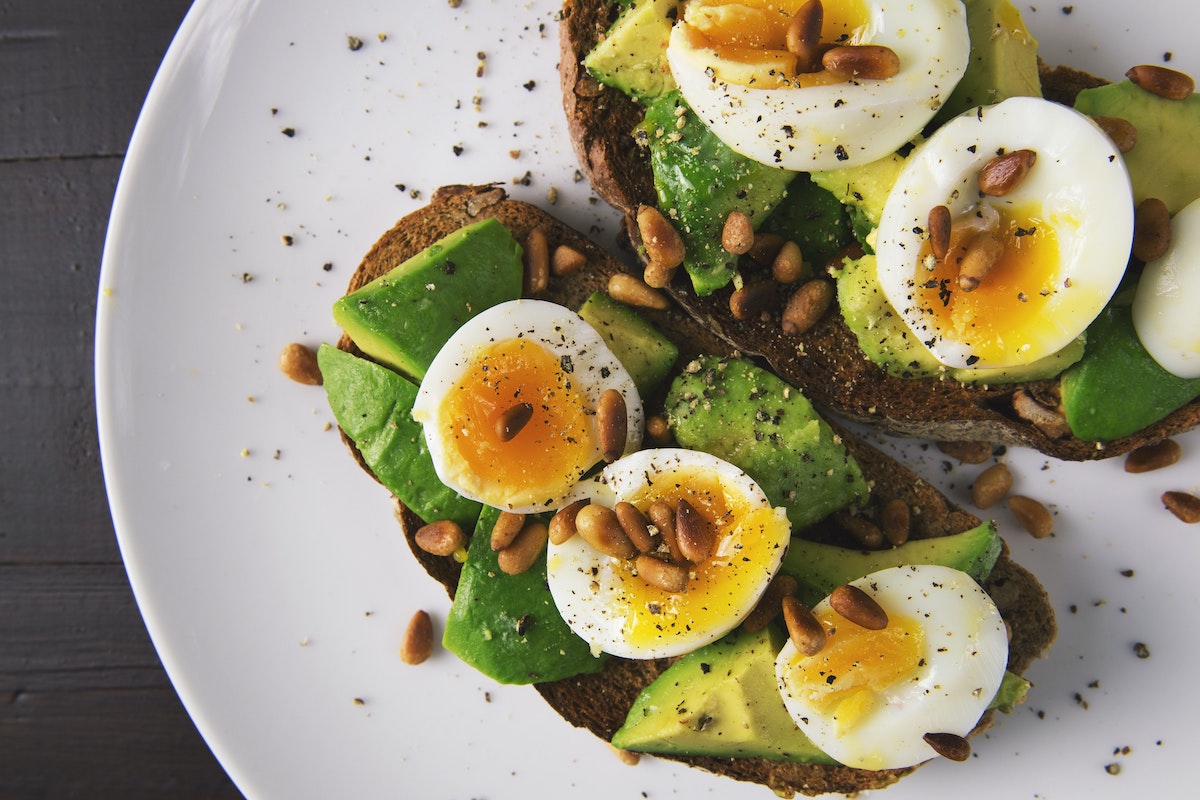 Two pieces of avocado toast topped with jammy eggs and roasted pine nuts on a white plate.