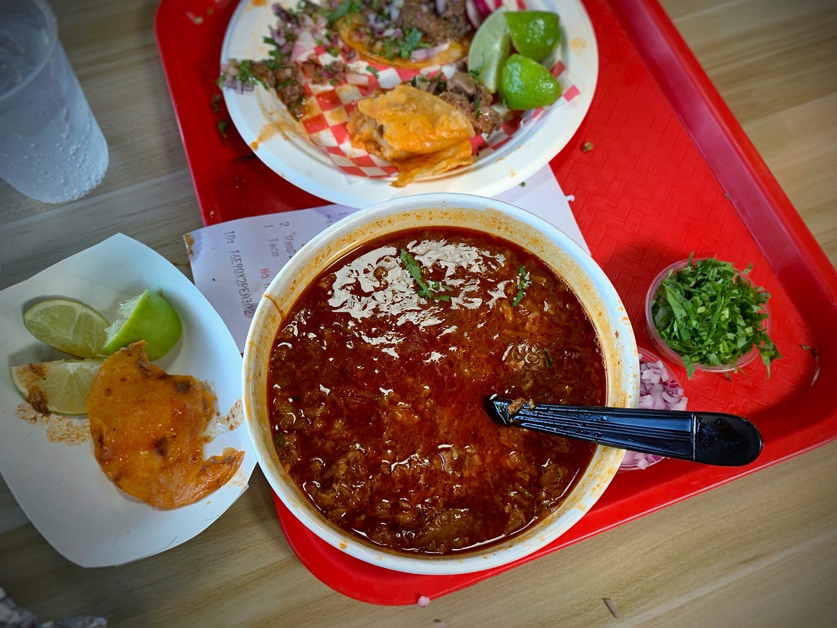 A bowl of hearty red soup next to a plate of tacos on a red tray.