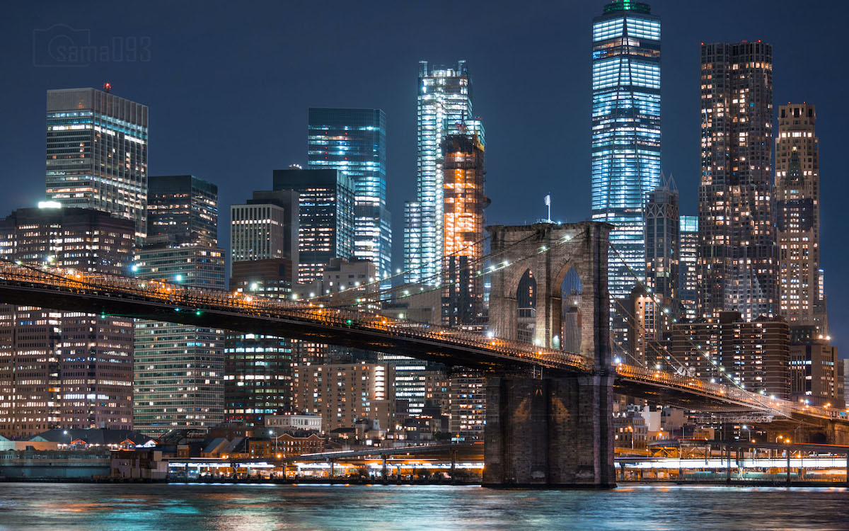 View of the Manhattan skyline and the Brooklyn Bridge lit up at night, taken from across a body of water. 