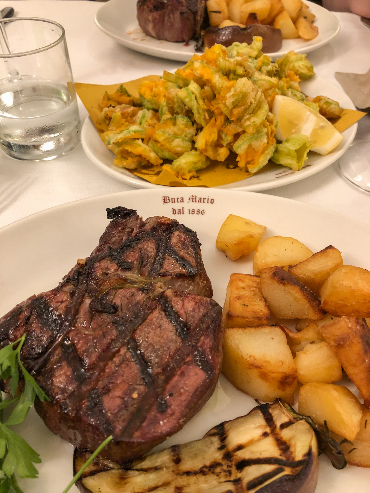 Grilled steak on a plate beside cubed potatoes, with other dishes visible on the table in the background