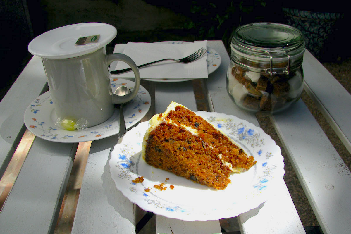 Slice of carrot cake next to a mug of tea and a glass jar of sugar cubes.