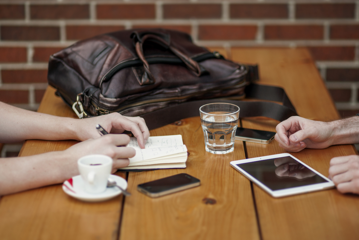 Wooden cafe table with a small espresso cup on a saucer, a small clear glass of water, a cell phone, a tablet, a notebook, and a backpack