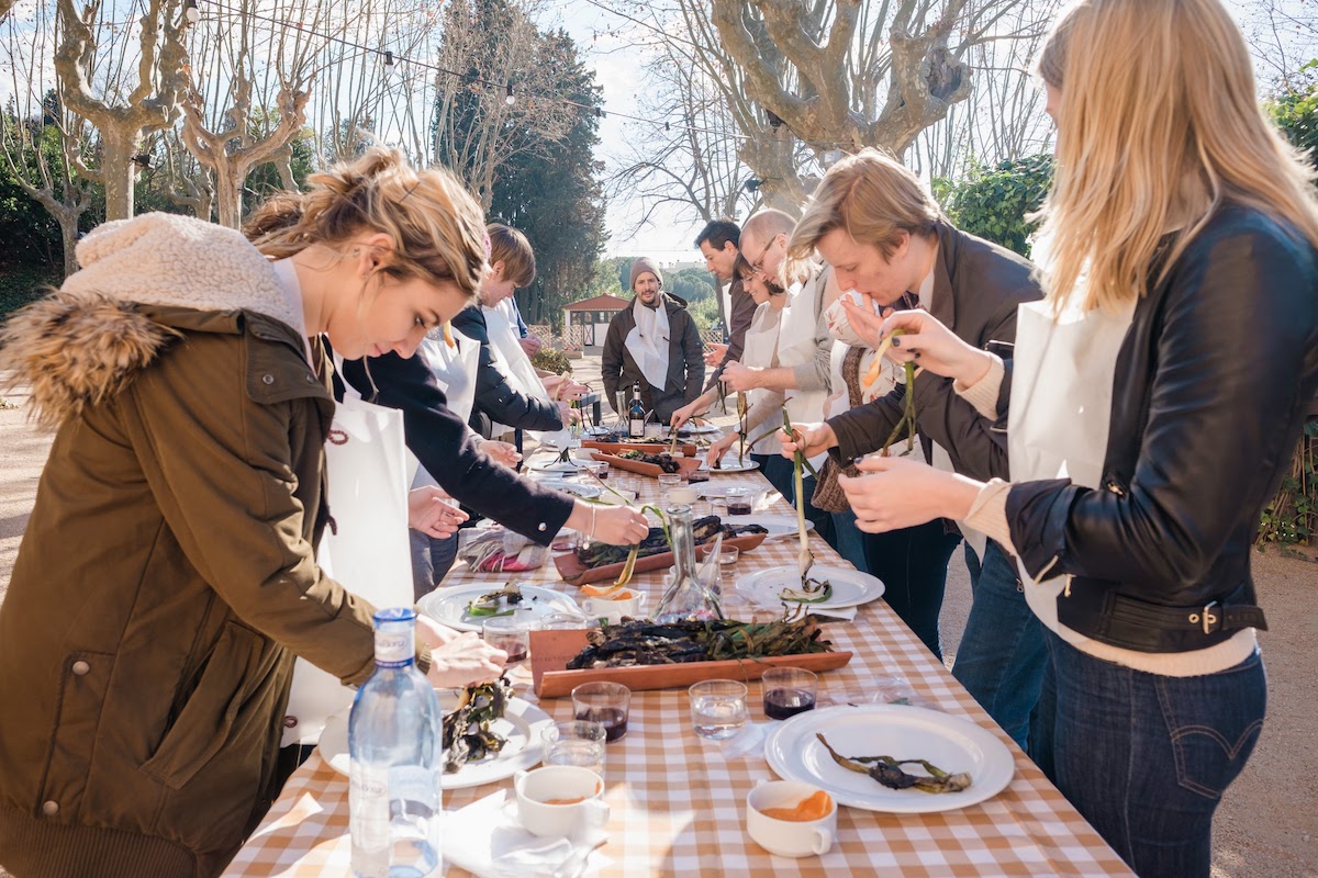 A group of people standing around a table outdoors eating grilled vegetables