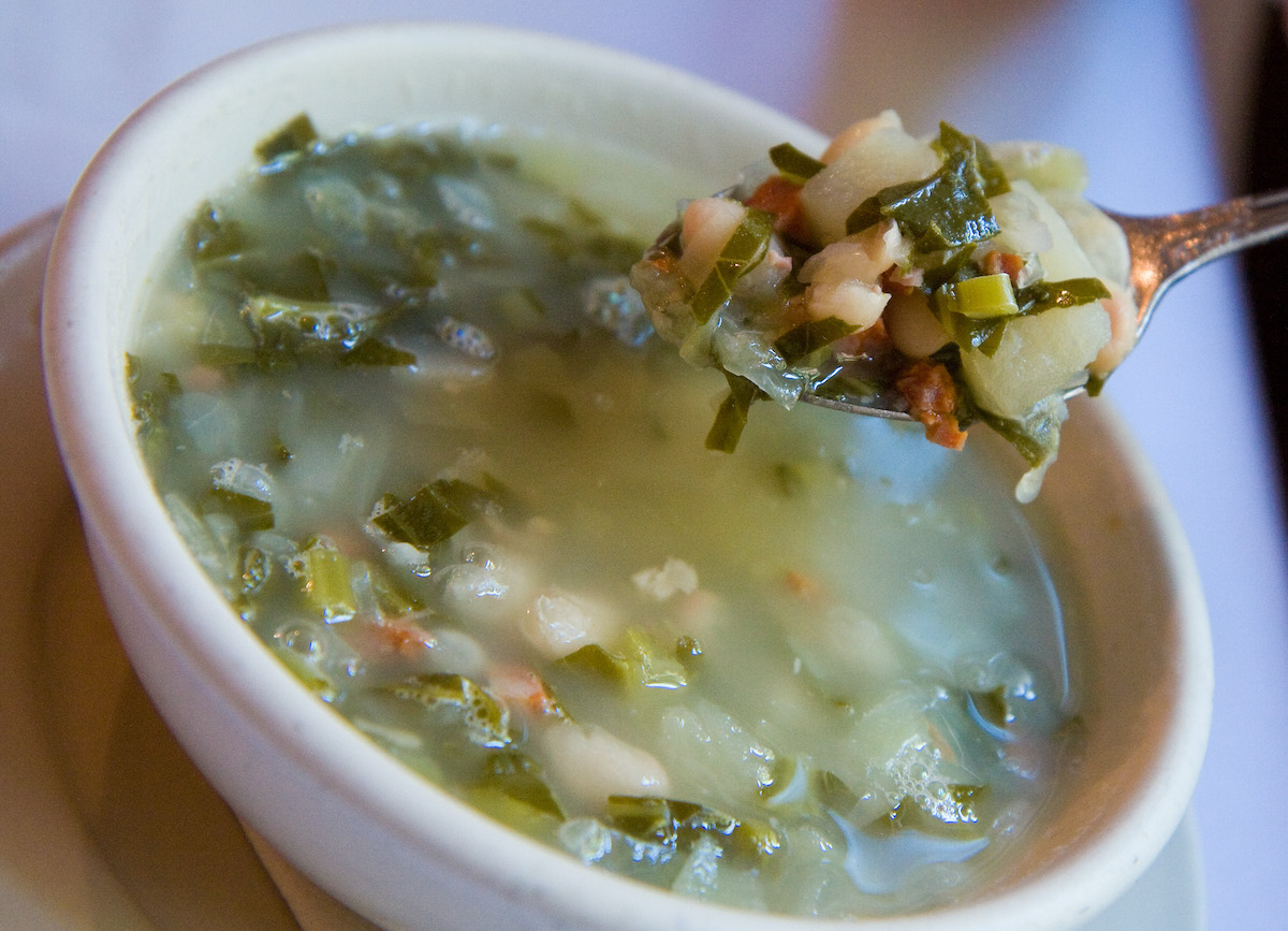 Close up of a bowl of Galician soup with greens, beans, and pork, with a spoonful being held by someone out of frame hovering above it.