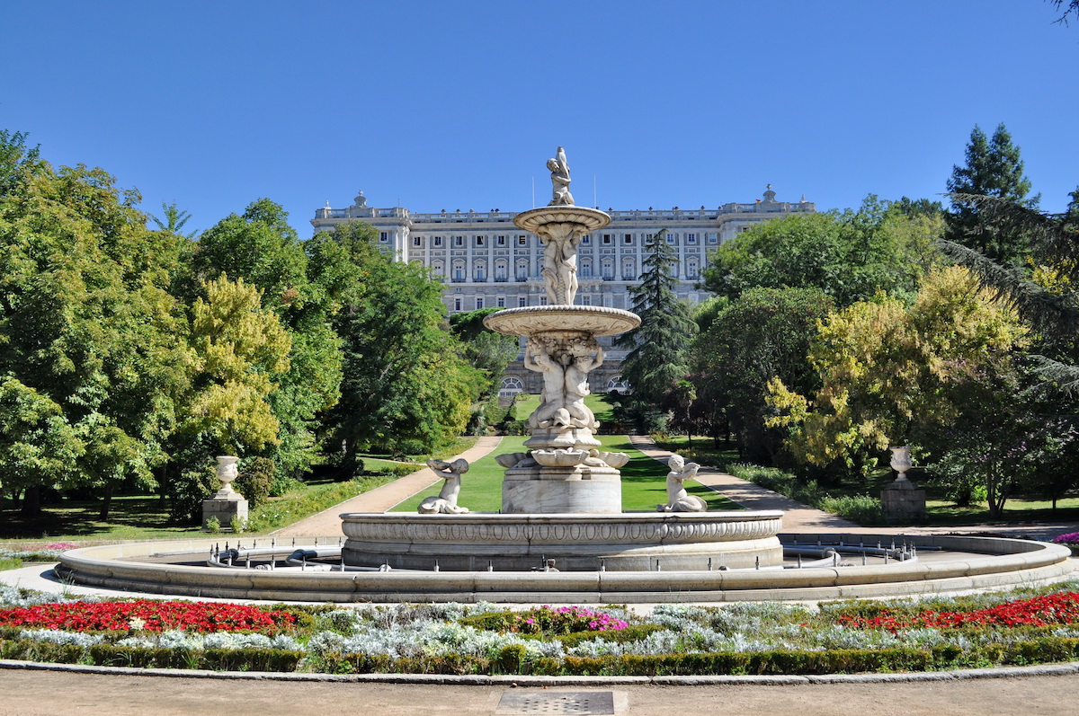 Large stone fountain surrounded by trees and flowers with a palatial building in the background.