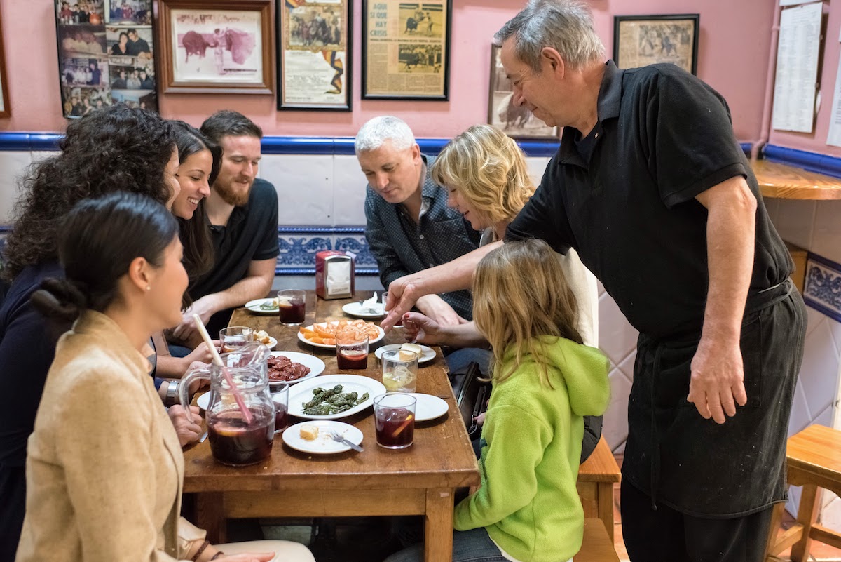 Family with kids at a restaurant in Madrid sitting around a small wooden table full of shared plates as a server places more food on the table.