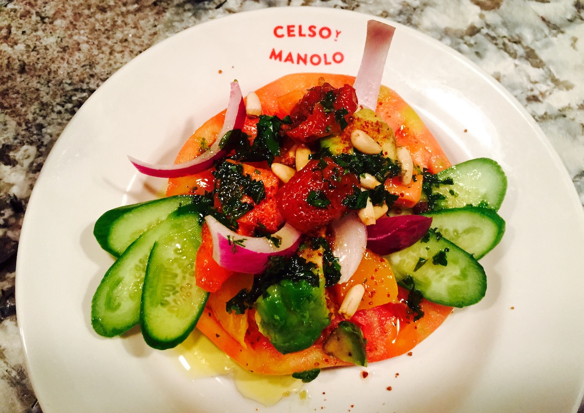 Vegetable dish with a base of tomato and garnished with red onion, cucumber, red peppers, and herbs, served on a white plate reading Celso y Manolo