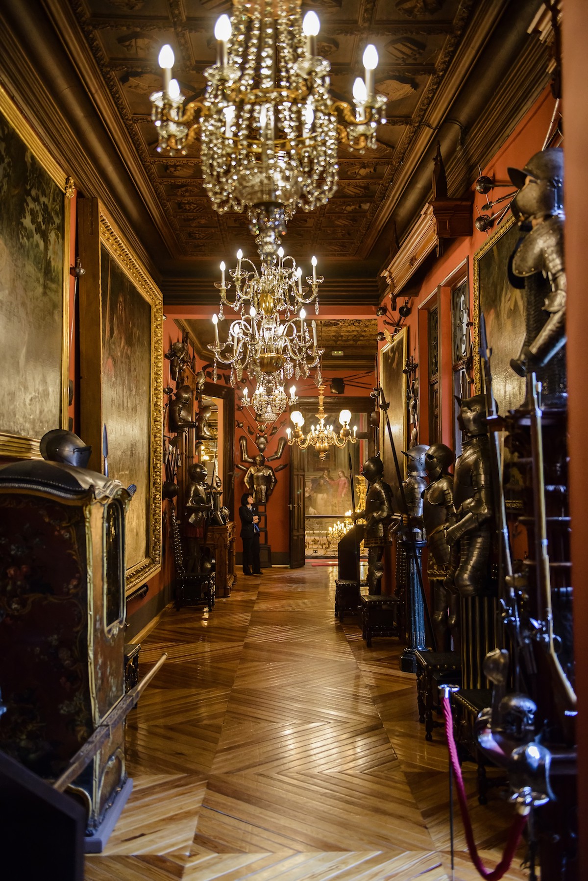 Long hallway lined with suits of armor with ornate chandeliers hanging overhead