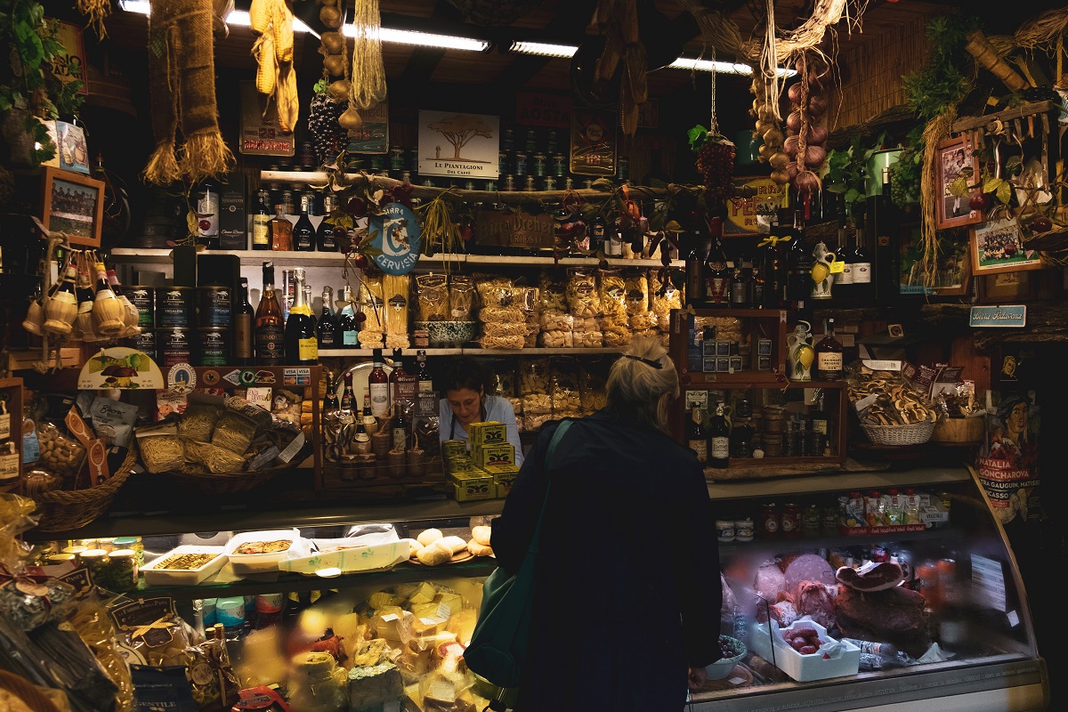 A middle-aged woman shops at a food stall packed with meats, cheese, breads, and packaged goods inside a market in Florence