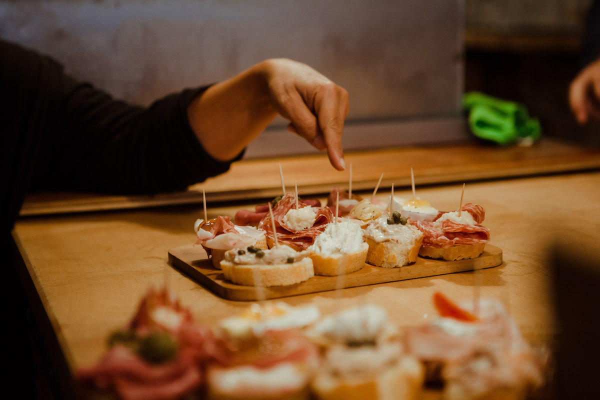 Small bites served on little pieces of bread placed atop a wooden board, with a person's hand pointing to them