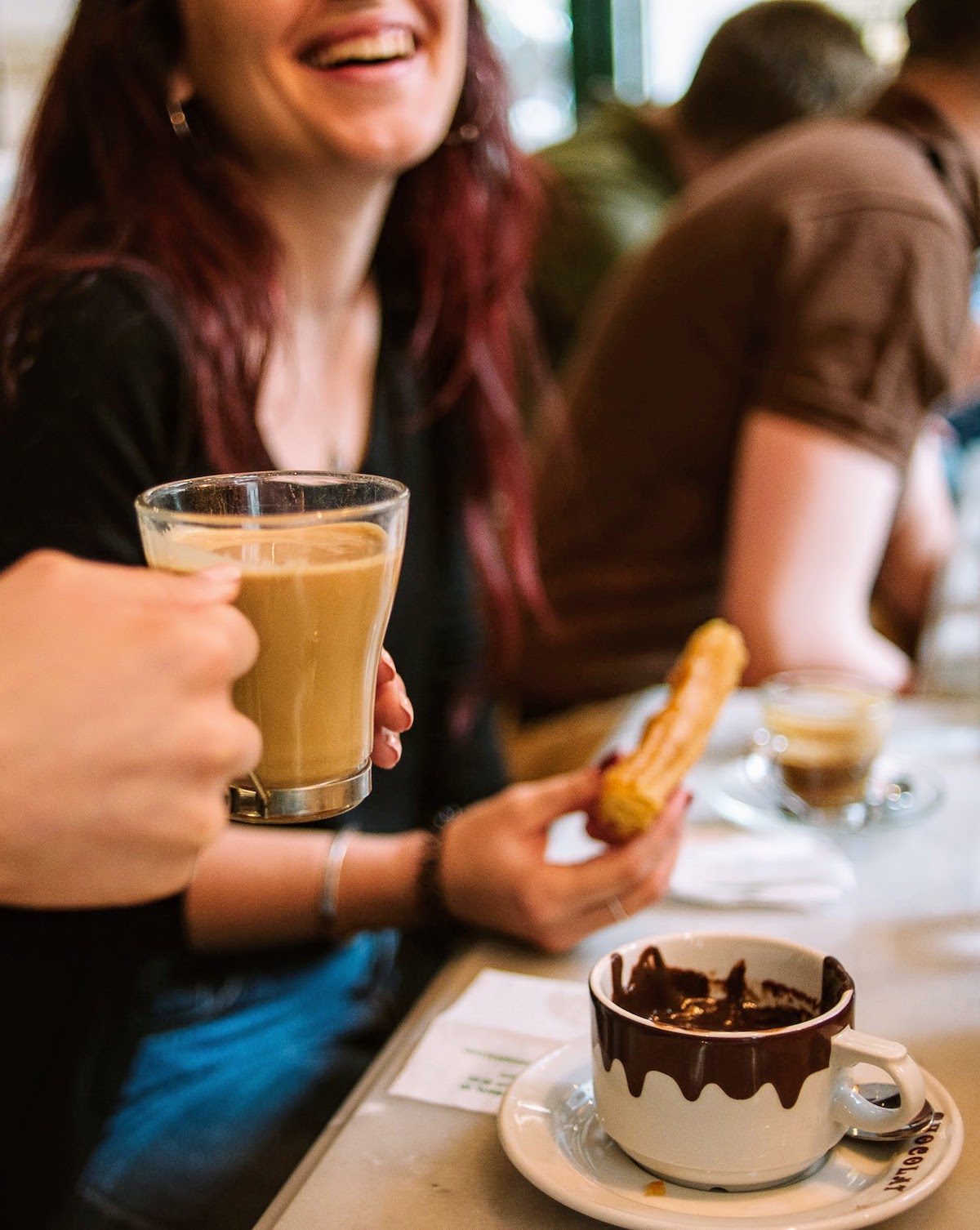 A person's hand holding a clear glass of coffee, with a woman laughing while eating churros and chocolate visible in the background
