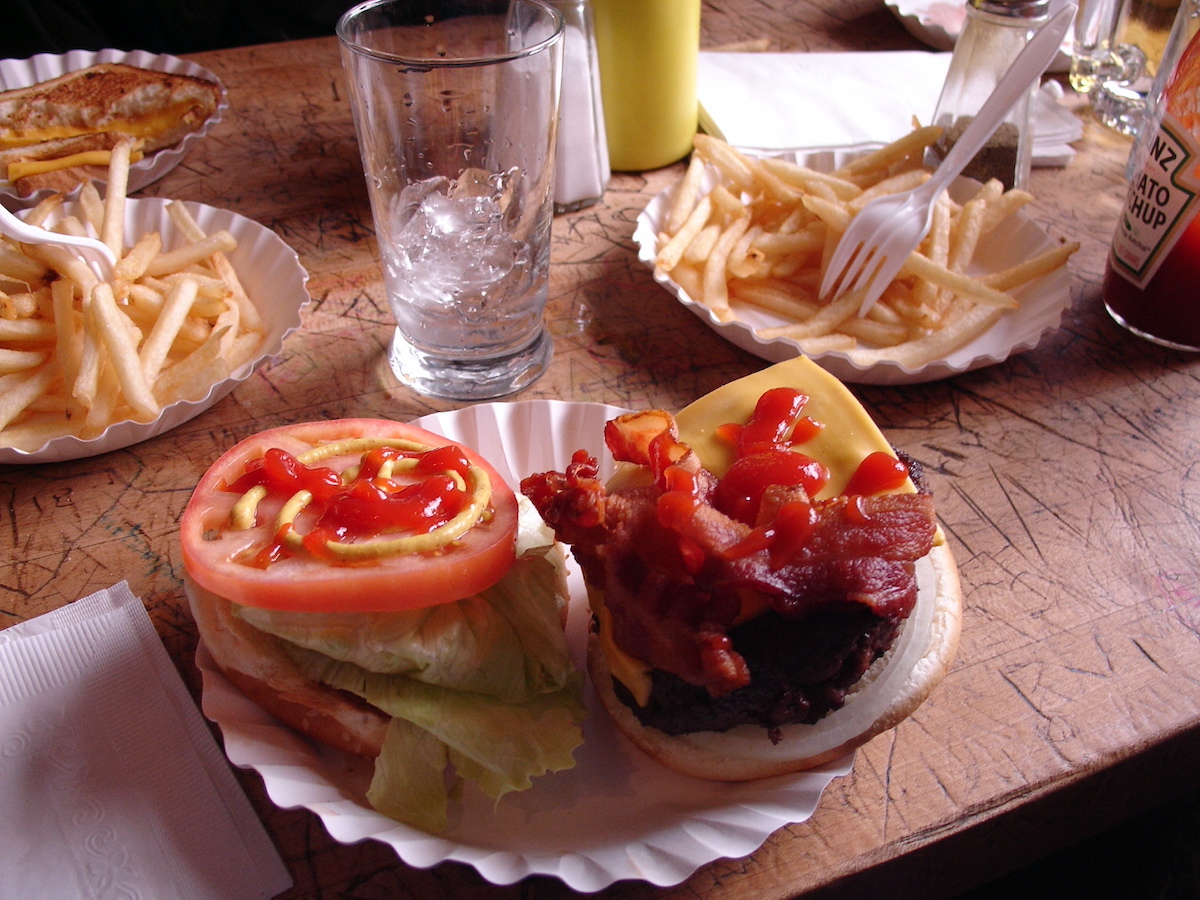 Open cheeseburger with lettuce, bacon, tomato, and ketchup served on a white paper plate.